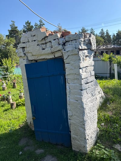 A weathered, partially collapsed stone structure with a bright blue wooden door stands outdoors in a grassy area with scattered potted plants and trees in the background. The structure's top is broken, revealing exposed brickwork and debris. A small, brass padlock hangs from the door's latch. The environment suggests a rural or semi-rural setting, possibly a garden or yard. The image is captured under a clear blue sky on a sunny day. The location is identified as Luka, Ukraine.