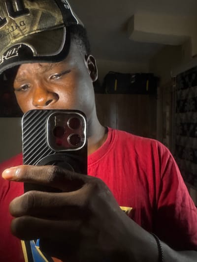 An image, appearing to be a reflection or mirror selfie, depicts a dark-skinned individual indoors in Ihiagwa, Nigeria. The subject, likely male, is shown from the chest up, wearing a red t-shirt and a camouflage-patterned baseball cap. They are holding a smartphone in their right hand, with the index finger extended. The phone's three-lens camera array and a distinctive textured, striped case are prominently visible. A black beaded bracelet adorns the person's left wrist. The individual's face is visible, looking towards the camera with a neutral expression.
The background is dimly lit, showcasing a light-colored wall or shelf on the left and a dark, rectangular object, possibly furniture, further back. A patterned surface, suggestive of a ceiling or wallpaper, is also present. Visible text on the baseball cap includes "DYNASTY" or a similar word on the front, and "SA" or "3A" on the side of the brim. A partial yellow and blue graphic design is discernable on the red t-shirt. The overall low-light conditions indicate an indoor setting, potentially during the evening or in a dimly lit room.