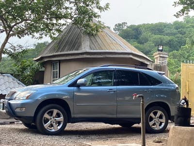 In Mararaba, Nigeria, a light blue Lexus RX SUV is parked on a gravel surface in front of a building with a distinctive conical, corrugated metal roof. To the left of the SUV, a large tree with green foliage extends over the scene. Behind the building, a dense green forest covers a hillside. A water tap with a red handle stands in front of the SUV. To the right of the SUV, a black bucket hangs on a yellow structure. The sky is overcast.