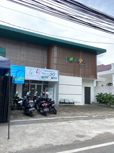 The image shows the exterior of a building located in Kecamatan Margahayu, Indonesia.  The building is a two-story structure with a green roof and wooden siding.  The front of the building features a sign that says "Konoha Clinic".  There are five motorcycles parked in front of the building.  A woman is featured on a promotional banner outside the clinic. The banner advertises a 50% discount on treatments.  Several power lines are strung above the building.  There is a white curb along the front of the building, and a small tree is visible in the foreground, to the right of the image.