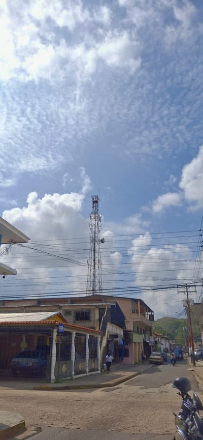 The image captures a street scene in Tinaquillo, Venezuela, under a partly cloudy sky. A prominent telecommunications tower stands tall in the mid-ground. In the foreground, a street is visible with houses and businesses lining its sides. A woman is walking away from the viewer on the sidewalk. A motorcycle is parked in the lower right portion of the frame. The environment appears to be a residential or commercial area of the city.