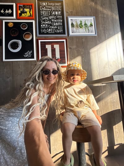 In an indoor setting with wood-paneled walls, a woman with long blonde dreadlocks and sunglasses smiles at the camera, holding a selfie stick. Beside her, a young child wearing a checkered hat and a light-colored t-shirt sits on a stool. The background features several framed pictures on the wall, including one with text and another with an illustration of the number 11. The lighting suggests it is daytime, with sunlight casting shadows. The overall scene appears to be a casual, possibly a cafe or restaurant, environment.