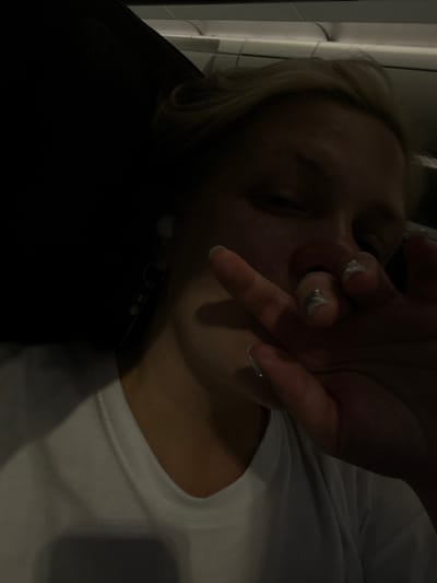 The image shows a close-up, low-light selfie of a young person with blonde hair, resting their face on their hand. They are wearing a white t-shirt. Their fingernails appear to have a metallic or glittery finish. A small, light-colored object, possibly an earring, is visible near their ear. The background is dark, with a hint of what appears to be the interior of a vehicle or a similar enclosed space, possibly indicating travel. The lighting is dim, casting shadows and obscuring finer details. The person's eyes are partially closed, and their expression is difficult to discern due to the lighting and hand placement. The overall impression is that of a candid, personal moment captured in low-light conditions, possibly during a journey. The location is stated as Roissy-en-France, France.