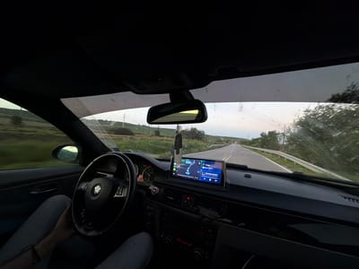 The image shows the interior of a car, specifically the dashboard, steering wheel, and part of the driver. The car is moving on a rural road, with green fields and some trees visible through the windshield. The GPS screen shows the car is 31 km away from Varna, Bulgaria. Based on this information, and the visible countryside, it is plausible that the car is in the vicinity of Boryana, Bulgaria, or on a road leading to or from it. The driver is holding the steering wheel, and wearing gray trousers. The car's dashboard has various controls and a navigation screen, and a decorative item hanging from the rearview mirror. The view from the windshield is slightly obscured by water marks. The light suggests it is either early morning or late evening.