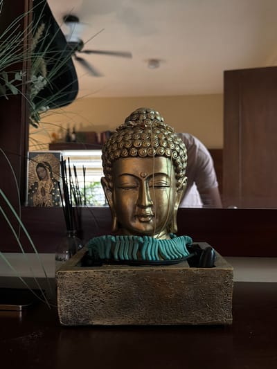 A gold Buddha head sculpture rests on a dark brown shelf. Turquoise elements accent the sculpture. Behind the sculpture, a mirror reflects the scene, showing a person in a white shirt and a ceiling fan. Plants and a religious icon are also visible in the reflection. A stone-like holder anchors the sculpture.