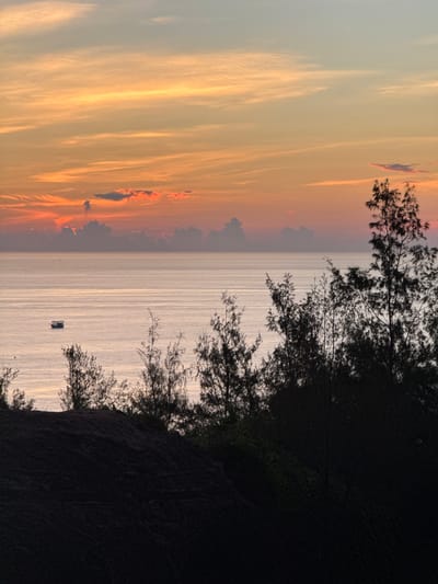 The image captures a serene seascape at dusk, with a warm, gradient sky of orange and pink hues stretching across the horizon. Thin, wispy clouds are scattered throughout the sky, reflecting the sunset's glow. In the distance, a silhouette of a small boat is visible on the calm, shimmering water. The foreground is dominated by the dark, silhouetted forms of trees and a sand dune, creating a stark contrast against the brighter sky and sea. The overall atmosphere is peaceful and tranquil, characteristic of a coastal sunset. The location is Lý Sơn Special Administrative Region, Vietnam.