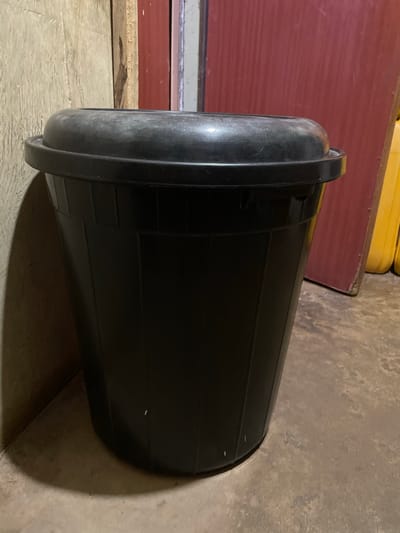 In Narayi, Nigeria, the image depicts a static indoor scene focused on a black plastic bin. The bin, positioned centrally, is cylindrical with a tapered body and vertical structural ribs. A matching black plastic lid, featuring a raised, domed center, is securely seated on the bin. Both the bin and its lid exhibit minor surface wear consistent with use.
To the immediate left of the bin, a textured, light-colored wall, possibly concrete or plaster, displays visible imperfections and a distinct curvilinear shadow cast by the bin. Further left and behind the bin, a vertical wooden element, likely a doorframe or support beam, is partially visible.
Behind the right side of the bin, a large, dark red vertical surface, consistent with a door or wall panel, shows signs of wear and possibly vertical grooves. In the far right background, partially obscured by the red surface, several yellow, rounded containers, possibly jerrycans, are visible, suggesting storage.
The bin rests on an uneven, multi-toned floor, appearing to be concrete or compacted earth, exhibiting varied texture and subtle debris. Ambient lighting casts shadows to the left and foreground of the bin, indicating a non-direct or somewhat dim light source. No individuals are present, and no actions or interactions are depicted. The overall environment appears utilitarian.