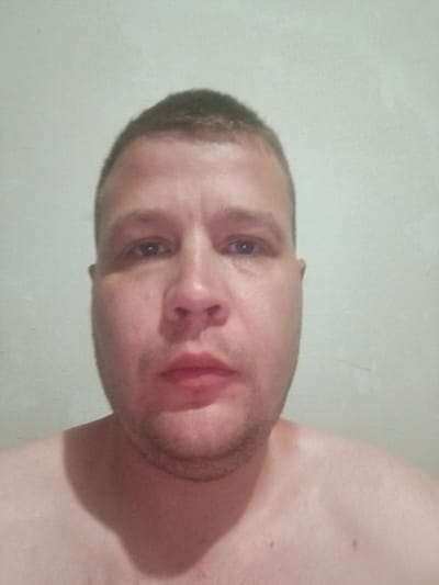 A close-up, head-and-shoulders, waist-up, chest-up, and shoulder-level selfie shows a fair-skinned adult male with a buzz cut, facing forward. The man has a full face, a prominent nose, and closed lips. His eyes are open and looking directly at the camera. He is shirtless, revealing his chest and shoulders. The background is a plain, light-colored wall. The image quality is moderate, with some graininess and soft focus. The lighting appears to be from the front. The man's skin tone is pinkish, suggesting he might have a flushed complexion or be fair-skinned. There are no discernible objects or other individuals in the frame. The environment appears to be indoors, likely a room with a plain wall. The image does not contain any visual elements that would place it specifically in Chirchik, Uzbekistan.
