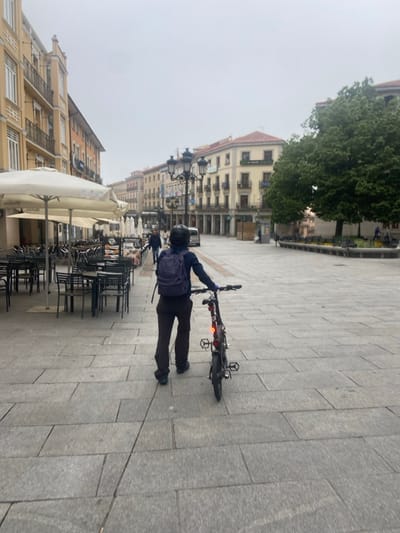 A person is walking on a paved square in the city of Segovia, Spain, near the Plaza Mayor (latitude: 40.948, longitude: -4.118). They are carrying a dark blue backpack and walking with a black bicycle. The square is mostly empty. In the background, there are buildings with various colors of paint. There is one large tree on the right. The square is mostly empty except for a few tables and chairs in the distance. There are a few other people walking around, and one person is sitting on a bench in the distance. The sky is gray and overcast. There is a light pole with street lamps in the middle of the square. The person appears to be in their late 20s or early 30s and is wearing a navy blue hoodie and black pants. The bicycle is black and silver. The square is mostly empty.