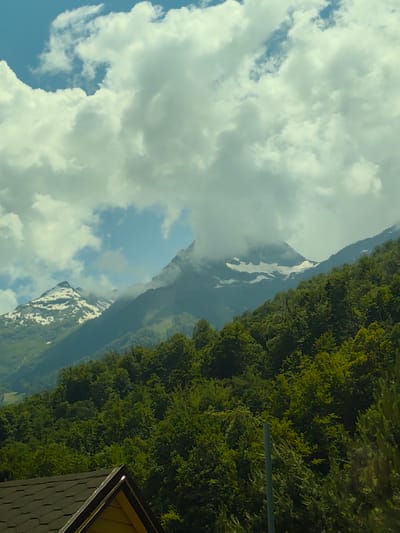 The image depicts a scenic mountain landscape under a partly cloudy sky. The foreground features a section of a dark-tiled roof with a visible yellow wall, suggesting a human-made structure. Behind the roof, lush green trees dominate the mid-ground, with their dense foliage creating a vibrant texture.

Further back, majestic mountains rise, their peaks partially shrouded in soft, white clouds. Some of the higher mountain slopes are dusted with snow, indicating a high altitude and potentially cooler temperatures despite the greenery below. The sky is a mix of bright blue patches and large, fluffy white clouds, suggesting daytime with variable weather conditions. The overall impression is one of natural beauty and tranquility. There is no discernible activity or event taking place, and no visible text. The scene likely represents a view from a hillside location in Estosadok, Russia.