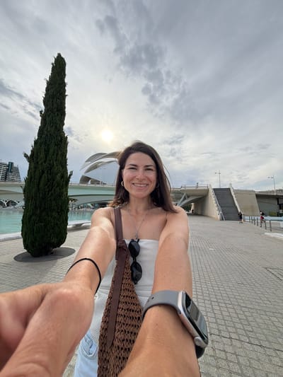 A woman takes a selfie in a city park in Valencia, Spain, with a tall, slender evergreen tree to her left and a futuristic architectural structure in the background. The woman is smiling and facing the camera, holding the phone with her right arm extended. She is wearing a strapless white top, denim shorts, and a brown woven shoulder bag. A silver smartwatch is visible on her left wrist. The ground is paved with light gray cobblestones, and there is a body of water to the left of the tree. The sky is cloudy with the sun peeking through. The architectural structure appears to be part of the City of Arts and Sciences, a complex of cultural and entertainment buildings.