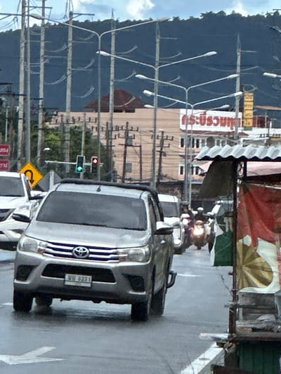 The image depicts a street scene in Chalong, Thailand. A silver SUV with a license plate reading "UU 6231" is the primary focus, positioned on the right side of the frame. The SUV appears to be in motion or recently stopped, with its headlights on.
Behind the SUV, a traffic light shows a green light for traffic moving horizontally across the image, and a red light for traffic moving vertically. A yellow road sign with a black arrow indicating a U-turn is visible below the traffic light.
Further in the background, there are buildings and numerous utility poles with electrical wires crisscrossing the scene. A large, dark blue structure, possibly a building or a shaded area, dominates the left side of the image. The sky is visible at the top left, with some clouds.
Several signs with Thai script are visible on a building on the left side, but the text is mostly unreadable due to the image quality and orientation. One sign appears to be red and white. A motorcycle with its headlight illuminated is also visible in the middle background, adding to the sense of an active street.
The overall impression is of a bright, daytime scene on a busy street. The perspective is angled, as if taken from a moving vehicle or a slightly elevated position. The lighting suggests it's daytime, and the weather appears to be clear. There are no clearly discernible people, and no overt emotional cues are present in the image. The setting is clearly urban and likely a commercial area given the signage and traffic.