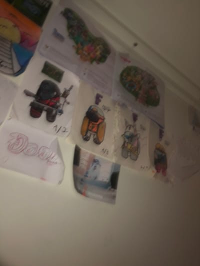 The image displays a wall with several drawings and artwork taped to it. The artwork consists of colorful illustrations, including what appears to be a character resembling an astronaut or a robot with a weapon, a bunny-like character, and several other abstract and patterned designs. One drawing in the lower left corner shows the word "Dami" written in a stylized font. The overall impression is of a child's decorated wall. The lighting is dim and somewhat blurry, suggesting the photo was taken in low light conditions or with a shaky hand. The location is not discernible from the image itself, and therefore it cannot be confirmed to be in L'Hospitalet de Llobregat, Spain.