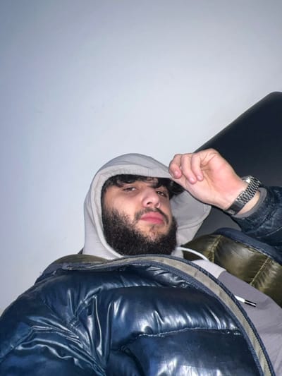 The image displays a close-up shot of a person, rotated approximately 90 degrees counter-clockwise. The subject is a male with dark, curly hair and a full beard and mustache. He is wearing a light-colored (possibly light grey or off-white) hooded sweatshirt, with the hood pulled up around his face. Over the hoodie, he has on a dark, shiny puffer jacket, which appears to be navy blue or black. A metallic watch with a link bracelet is visible on his left wrist.
The person's head is tilted, and he is looking directly towards the camera with a neutral or slightly contemplative expression. His lips are slightly parted. His left hand, with fingers curled, is raised and positioned near the edge of his hood.
The background is simple, consisting primarily of a plain, light-colored wall (white or light grey) occupying the majority of the left side of the frame. To the upper left, an angled dark surface, possibly a part of a piece of furniture like a chair or sofa, is visible behind him. The lighting appears even and soft, suggesting an indoor setting. No text is visible in the image.