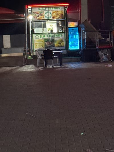 The image depicts a roadside smoothie stand in Kurunduma, Nigeria. The stand, named "S.A.F BLENDS", has an illuminated sign and displays images of its offerings. A brightly lit cooler with the name "NEXUS" on top is positioned nearby. Two people are seated at a small table in front of the stand. The stand is situated on a paved surface.