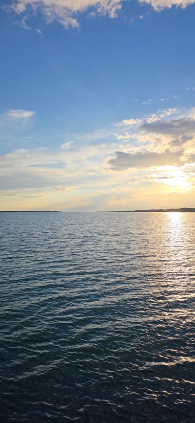 This is a vertical, eye-level shot of a calm body of water meeting the sky at sunset. The image is split diagonally, with the left side dominated by a bright blue sky filled with wispy white and grey clouds, some illuminated by the sun. The sun itself is a bright, hazy orb positioned just above the horizon on the right side of the frame, casting a warm, golden light that blends into the sky and reflects off the water.
The right side of the image is the water, likely a lake or sea, reflecting the sunset. The water's surface is rippled, creating a textured, shimmering effect as the sunlight catches the waves. The horizon line is clearly visible, separating the sky and water, with a faint, dark line of land visible in the distance. The overall mood is serene and peaceful, conveying the beauty of a natural landscape at dusk. The lighting suggests it is late afternoon or early evening. There is no visible text in the image.