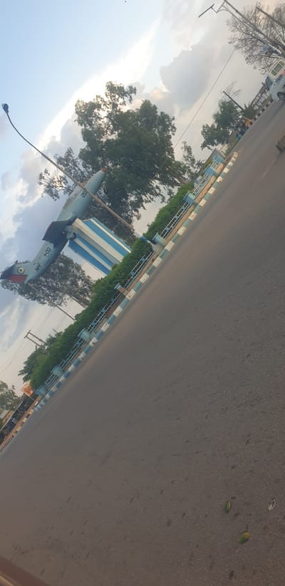 A light blue jet aircraft is displayed on a pedestal in the city of Barakin Lerawi, Nigeria. The aircraft is positioned at an upward angle, with its nose pointing towards the sky. It is mounted on a cylindrical pedestal that is painted with alternating blue and white stripes. The pedestal is surrounded by a low green hedge and a decorative fence with blue and white striped borders. The aircraft has markings on its tail, including a circular insignia with red, green, and white colors. The registration number "NAF 420" is visible on the fuselage. The scene is captured on a road, with asphalt visible in the foreground and extending into the background. Trees with dense green foliage are scattered throughout the scene, and utility poles with wires are visible in the distance. The sky is partly cloudy, with patches of sunlight breaking through. In the background, a white vehicle and some buildings are partially visible, suggesting an urban or semi-urban setting. The overall impression is that of a monument or display, possibly located near an airport or military installation.