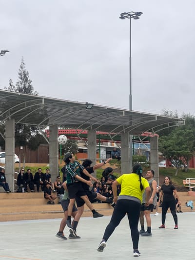 In Arica, Chile, a group of people are playing a game of futsal on an outdoor court under an overcast sky. The court appears to be part of a recreational park, with a covered seating area and bleachers visible in the background. Several individuals are actively engaged in the game, with two men in mid-air, one about to head a soccer ball, and another in pursuit. Other players are positioned on the court, some observing the game and others ready to join in. Spectators are seated on the bleachers, watching the match. The environment is characterized by urban infrastructure, including a tall light pole and a covered structure with concrete pillars, suggesting a well-maintained public space. The overall scene depicts a casual sporting event in a community setting.