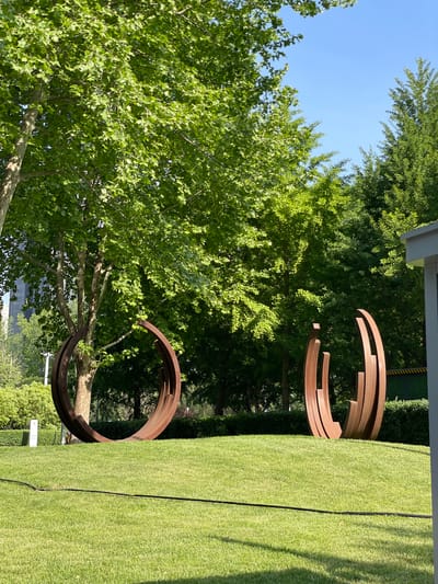 The image depicts an outdoor scene in Chaoyang District, China, featuring two large metal sculptures on a grassy lawn surrounded by trees. One sculpture is composed of several concentric, incomplete circles stacked vertically, while the other consists of a single large, incomplete circle. The grass appears well-maintained and vibrant green. The trees provide a backdrop of dense foliage, partially obscuring the sky, which is visible as a clear blue in the upper left corner of the image. The sculptures are made of a rusty-colored metal, contrasting with the green of the lawn and trees. The scene is captured in bright daylight.