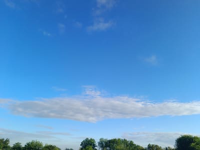 The image captures a broad expanse of a clear blue sky, dotted with scattered white clouds. A large, elongated cloud bank stretches horizontally across the middle ground of the frame. Below this, a line of lush green trees forms the lower boundary of the image, silhouetted against the sky. The scene suggests an outdoor environment, likely a park or rural area on a bright day. The location is identified as Volodymyrivske, Ukraine. There are no people or discernible objects other than natural elements present in the scene.
