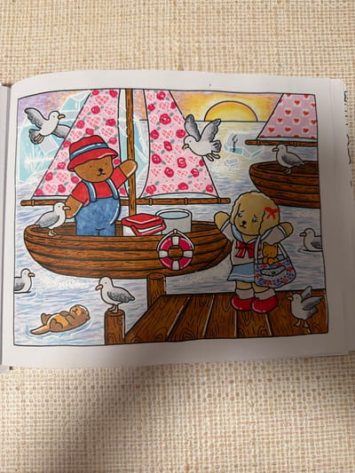 The image depicts a colorful illustration of a seaside scene. In the foreground, a wooden dock extends into the water. A yellow dog wearing a sailor outfit stands on the dock, holding a purse containing a small brown teddy bear. Several seagulls are scattered around the dock and in the water. To the left, a brown bear in a red hat and blue overalls stands in a wooden boat with a red and white striped sail adorned with pink roses. Another boat with a pink sail decorated with red hearts is visible in the background, near a sunset over the water. A sea otter floats peacefully in the water near the dock. The image is rendered in a whimsical, cartoonish style, suggesting it is from a children's book or coloring page. The specific location in Boca Raton, United States of America, cannot be determined from the provided image.