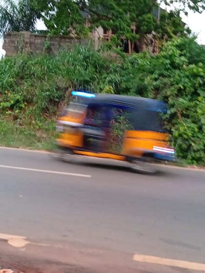 A blurry photograph shows a moving vehicle, possibly an auto rickshaw, on a road in Ogidi, Nigeria. The vehicle appears to be yellow and blue. Green foliage and a partially constructed structure are visible in the background. The road is grey and has a white line marker. The blur indicates motion.