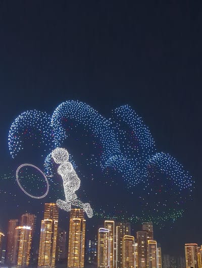 The image captures a nighttime cityscape in Jiangbeicheng Subdistrict, China, characterized by a drone light show above illuminated skyscrapers. The drone display forms a large, abstract shape resembling wings or a cloud, composed of thousands of tiny white and colored lights against the dark sky. In the foreground of the sky, a figure made of white lights appears to be a stylized human, possibly depicted in a playful or athletic pose, with a ring of lights in front of it. Below this aerial spectacle, a dense collection of modern high-rise buildings with glowing windows lights up the urban landscape. The lighting on the buildings is predominantly orange and yellow, with some outlines picked out in white or blue, indicating an active and vibrant city at night. The overall scene is one of technological spectacle and urban development.