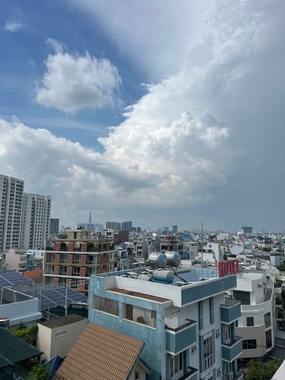 The image is a high-angle, panoramic view of a dense urban cityscape in Thủ Đức, Vietnam, under a partially cloudy sky. The composition is divided, with the left side dominated by dramatic clouds and blue sky, and the right side showcasing a sprawling collection of buildings.
The cityscape consists of a mix of architectural styles and building heights. Numerous multi-story residential and commercial buildings are visible, many with light-colored facades and varying window designs. Some buildings feature balconies, while others are more utilitarian. Closer to the foreground on the right, a building with a bright red sign displaying the word "LUCH" is prominent. Below this, more buildings with light blue and grey accents are visible, some with tiled roofs. In the middle ground, solar panels are installed on several rooftops, suggesting an effort towards sustainable energy. Further in the distance, a hazy skyline of taller buildings, including what appears to be a skyscraper, can be seen.
The weather appears to be dynamic, with large, fluffy white clouds contrasting against patches of clear blue sky. The lighting suggests it is daytime, possibly late morning or early afternoon, with the sun illuminating the buildings and clouds.
No people are clearly discernible in the image. The scene is one of urban development and daily life, with the focus on the built environment and the atmospheric conditions. The overall impression is that of a bustling city with ongoing construction and modernization.