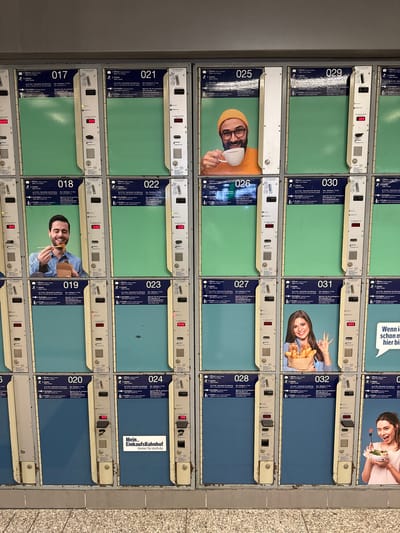 This is a medium shot, eye-level, outdoor photograph taken in Duisburg, Germany, featuring a row of automated luggage lockers. The lockers are arranged in a grid of two rows and multiple columns, with each locker identified by a number. The lockers are predominantly green and blue, with beige and silver metallic accents. Each locker has a digital display and operational buttons for opening, payment, and closing.
Several lockers display images of people. Locker 017 has a green front. Locker 018 shows a man in a blue shirt, smiling and eating from a takeout container. Locker 019 has a green front. Locker 020 has a blue front. Locker 021 has a green front and shows a man wearing a yellow beanie and holding a cup of coffee. Locker 022 has a green front. Locker 023 has a blue front. Locker 024 has a blue front and a sign that reads "Mein_Einkaufsbahnhof Immer für mich da." Locker 025 has a green front. Locker 026 has a green front. Locker 027 has a blue front. Locker 028 has a blue front. Locker 029 has a green front with a red digital display showing "3.00". Locker 030 has a green front. Locker 031 has a blue front and shows a woman holding a bag of bread. Locker 032 has a blue front. To the right of locker 032, a portion of a locker shows a woman in a pink shirt, winking and eating a salad with a fork. Above the lockers, the wall is a light gray color, and a section of a sign is visible on the right side, reading "Wenn ich schon mal hier bin". The floor in front of the lockers is a textured, light brown material. The overall scene is clean and functional.