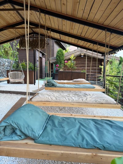A covered outdoor area features several wooden swings with mattresses and pillows, set on a gravel ground. The swings are arranged in a row and are suspended by ropes from a wooden ceiling. In the background, there are wooden structures resembling small cabins and lush green trees. A stone wall and a wicker chair are also visible. The overall scene suggests a serene and natural outdoor space.
