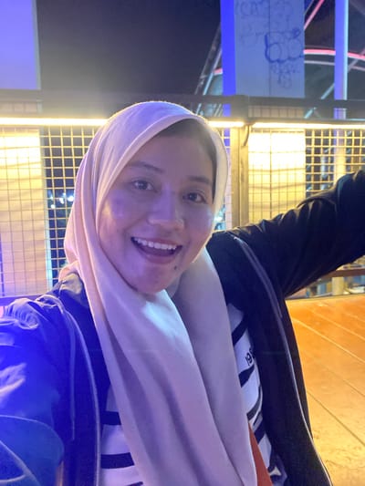The image is a selfie taken at night in Banjarbaru, Indonesia, likely near a Ferris wheel or amusement park attraction, given the background structures with blue and pink lights and graffiti. The subject, a woman wearing a light-colored hijab and a dark jacket over a striped shirt, smiles broadly, her teeth visible. Her right arm is extended out of frame, suggesting she is holding the camera or posing. The lighting is artificial and casts a purple hue on her face. The foreground shows a wooden floor, likely part of an outdoor seating area or walkway.