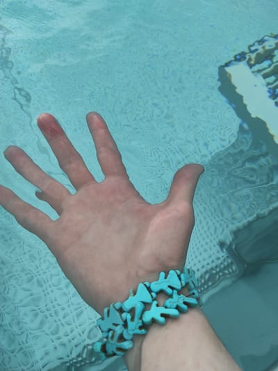 A close-up, high-angle shot shows a person's left hand submerged in clear, light blue water. The hand is open, with fingers spread and pointing towards the left side of the frame. The skin of the hand appears slightly reddened and is partially obscured by the rippling water, which refracts light and creates a mosaic-like pattern on the pool's surface. A turquoise bracelet, composed of interlocking, coral-shaped pieces, is worn on the wrist. The bracelet stands out against the skin tone and the watery background. The scene suggests the person is in a swimming pool or a similar body of water, possibly outdoors given the bright lighting and the presence of reflections from above. The water is calm, with only slight disturbances from the hand's movement. There is no visible text in the image.
