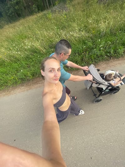 A high-angle, wide shot captures a family on a paved path in a rural area, possibly near Podshivalovo, Russia, with a grassy field and trees to the left. A man in a teal t-shirt and burgundy shorts pushes a gray stroller, in which a baby wearing a white hat lies. To the left of the stroller, a woman in a dark grey sports bra and grey sweatpants, also facing the camera, is taking a selfie. She has blonde hair pulled back and is wearing white sneakers. The scene appears to be during the daytime, with natural light illuminating the subjects and the surrounding greenery.