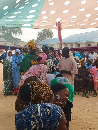 This outdoor image depicts a lively social gathering in Doma, Nigeria, under a large, temporary canopy. The canopy is made of fabric, featuring light green and beige sections with white polka dots, and is tied back in places.
Numerous people are present, a mix of adults, with many women dressed in colorful traditional African attire, including various head coverings and patterned dresses. In the center, a man in a light peach-colored tunic stands, appearing to interact with a woman who is bending forward in front of him. Other individuals surround them, observing the activity. Many faces show engagement, with some people smiling or laughing, particularly a woman in the foreground on the right wearing a black headscarf and distinct circular earrings. To the left, a group of women stands in what appears to be a line, some wearing hijabs.
In the background, additional attendees are seated on chairs. The ground beneath is bare earth or sand. Simple, single-story buildings with light-colored walls and corrugated metal roofs are visible in the distance, interspersed with trees, indicating a rural or semi-urban setting. The natural light suggests it is daytime, with no harsh shadows implying midday, possibly morning or late afternoon. The overall atmosphere appears communal and celebratory. There is no visible text in the image.