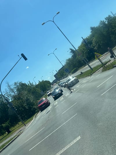 The image depicts a sunny day in Pilsen, Czechia, on a multi-lane road with a clear blue sky. The road has white lane markings and a pedestrian crossing. Several cars are visible, including a red Skoda and a white SUV, some of which are in traffic. A person with crutches is crossing the road. Trees line the right side of the road, and streetlights are present. A traffic sign with a yellow triangle and a black symbol is also visible on the right. In the distance, buildings and more trees can be seen.
