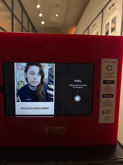 This image shows a red photo booth, likely a "one-cut" photo booth, in what appears to be a public space. The photo booth has a screen displaying a woman's selfie, taken in front of a backdrop with Korean text and images of food, possibly kim chi. The woman has light skin, blue eyes, and brown hair, and is wearing a dark blue turtleneck with white stripes. She is holding a smartphone with a snake-patterned case in her right hand, taking the selfie.
To the right of the screen, there's a set of instructions in Korean and English. The English text reads "Hello, Please press the button for the shoot". Below this, there are buttons labeled "Print" and "Reshoot". Further down, there's a sticker with a hand pointing and text in Korean that translates to "Stickers will come out with the photo!".
The text on the bottom of the screen reads "MUSEUM KIMCHIKAN". The text above the screen on the right side, under the heading "원픽스사용법" (likely "One-pic Usage"), provides instructions for using the photo booth.
The background of the image shows a dimly lit interior with a ceiling with lights and a reflective surface. The overall impression is that this photo booth is located in a museum or an attraction related to Korean culture, specifically kim chi, within Seoul, South Korea. The "MUSEUM KIMCHIKAN" branding strongly suggests this location.
