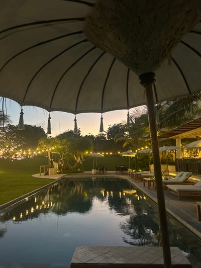 This is a medium shot photograph taken at dusk, likely at a resort or hotel, located in Kediri, Indonesia. The scene is centered around a swimming pool, with lounge chairs and umbrellas positioned on a wooden deck adjacent to it. The pool water reflects the surrounding lights and palm trees. In the foreground, a large umbrella partially obscures the view, with decorative tassels hanging from its edge. The background features lush greenery, trees adorned with string lights, and a hint of buildings with awnings. The sky displays a gradient of warm sunset colors. Two individuals are visible near a table under one of the lit trees, one appearing to be interacting with a counter or service area. The overall atmosphere is tranquil and inviting, suggesting an evening leisure setting.