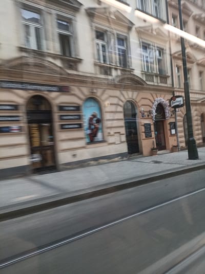 The image shows a street scene in Prague, Czechia, with a blurred effect suggesting movement, possibly from a vehicle. The foreground displays tram tracks and a road, with a sidewalk adjacent to a building facade. The building, rendered in muted tones of beige and cream, features architectural details like columns, arches, and decorative cornices. Several windows with white frames are visible on the upper floors.
On the ground floor, there are shops or establishments with arched doorways. One doorway is dark and appears to be an entrance, with horizontal slatted doors. Another entrance is decorated with a floral arch and leads to a establishment with a sign that appears to say "Pilsner Urquell". A black sign with white text, possibly a menu, is mounted on the wall to the left of this entrance. Further to the left, a large arched window or display area features a depiction of two people in silhouette against a light blue background. Above the storefronts, horizontal dark rectangular signs with white lettering are affixed to the building.
A street lamp post, black and tall, stands on the sidewalk, to the right of the building. Two potted plants with dark soil and green foliage are placed on the sidewalk near the entrance decorated with the floral arch. The street and sidewalk are paved with greyish stones. The overall impression is of a historical European city street.