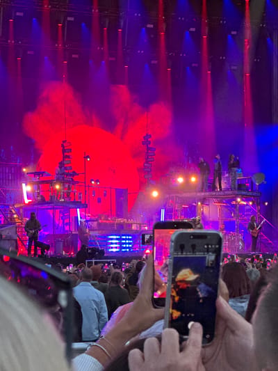 The image depicts a large outdoor concert at night, illuminated by vibrant red and blue stage lights. In the foreground, several people are holding up smartphones to record or photograph the performance. One phone in the center shows a close-up of a performer in a hat. The stage itself is multi-leveled with several musicians visible. On a higher platform, one individual is operating what appears to be musical equipment. On a lower level, a guitarist is standing and playing. Smoke or haze adds to the atmospheric lighting. The overall environment suggests a lively and energetic music event. The location is identified as the city of Glasgow, United Kingdom.