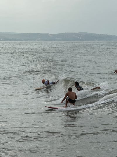 Four individuals are surfing on a choppy ocean under an overcast sky. The middle ground features three surfers actively riding waves. In the foreground, one surfer is positioned on a red surfboard, crouched low and riding a wave. To the left and slightly behind, another surfer lies prone on a light-colored surfboard. Further back and to the right, a third surfer is partially submerged in a wave, with only their head and shoulders visible. A fourth individual, further in the distance on the right, is partially visible, also in the water. The background consists of a hazy shoreline with hills and vegetation. This scene is likely located in the waters off the coast of Phan Thiết, Vietnam, a region known for its surfing conditions.