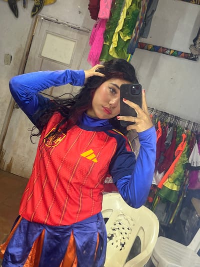 In this media file, a young woman is captured in a mirror selfie. She is wearing a red jersey with yellow stripes and an emblem that appears to be a coat of arms, over a blue long-sleeved shirt. Her lower body is covered by blue fabric that transitions into orange. Her hair is dark and wavy, and she has makeup on, including prominent eyeliner. Her right hand is holding a black smartphone, with a silver ring on her ring finger and red nail polish. She is touching her hair with her left hand.
The setting appears to be a room filled with various colorful fabrics and costumes, possibly a workshop or a backstage area. There are racks of clothing and piles of material in vibrant colors like green, yellow, pink, and red. In the background, white plastic chairs are visible, one of which is partially visible to the right of the frame. The lighting suggests it could be daytime, but the indoor setting with artificial light is also possible. The overall mood seems casual and perhaps playful, given the nature of the selfie and the colorful environment. There are no discernible weather cues. Location cues are limited to the background items which do not specifically identify Porlamar, Venezuela. There is no visible text in the image.