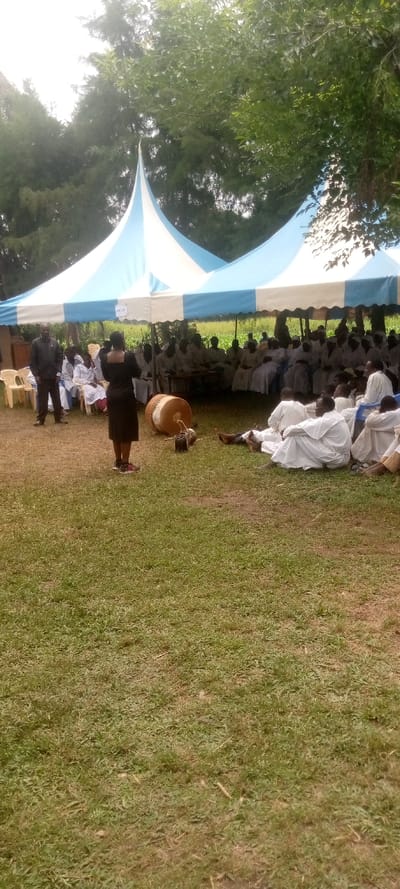 The image shows a gathering of people under two white and blue striped tents in a grassy field. The people are sitting in rows facing the camera, and a few individuals are standing in the foreground. A woman in black is standing in the middle of the frame, facing the camera. There is a large drum lying on the ground in front of the people. The location is a grassy field in Burundu, Kenya, which is located at the coordinates 0.459 latitude and 34.776 longitude. There are trees in the background.