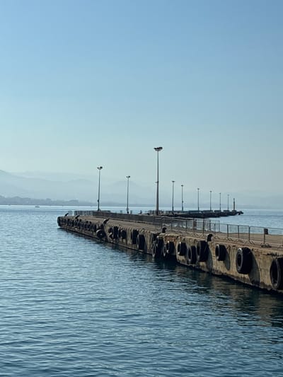 This is a photograph of a pier extending into a body of water. The pier is lined with what appear to be tires, likely used as fenders. Several lampposts are also visible along the pier, suggesting it's a place for activity, possibly fishing or recreation. The water is calm with gentle ripples. In the background, hazy mountains are visible, with a clear, pale blue sky above. The overall impression is one of a serene, early morning or late afternoon scene. There are no people or visible text in the image. The location is identified as Alanya, Turkey, which aligns with the coastal landscape. The lighting suggests it is either early morning or late afternoon.