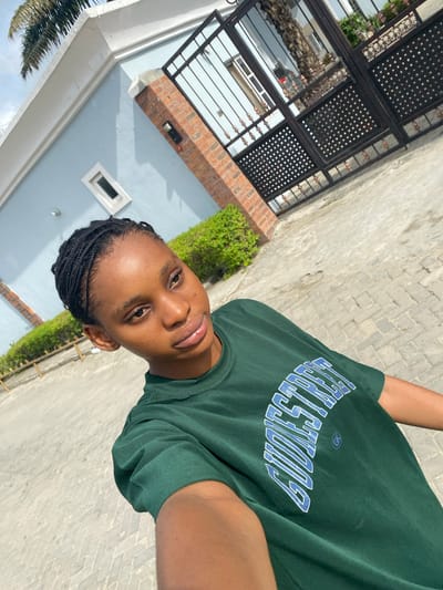 A person with braided hair is wearing a green shirt with blue lettering. They are standing outside near a building and pavement. The building is light blue with a brick detail. There is greenery present. The location is possibly Iranla, Nigeria.