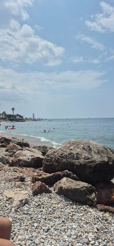 The image depicts a rocky beach in Paleo Faliro, Greece, with people swimming and relaxing in the calm, blue sea under a partly cloudy sky. In the foreground, large, weathered rocks and smooth pebbles cover the shoreline. The middle ground shows several individuals enjoying the water, some on inflatable rings. In the distance, a linear cityscape with buildings, including a prominent skyscraper, lines the coast. Palm trees are visible along the shore, and a few boats are scattered across the horizon. The overall scene is one of a typical summer day at a Mediterranean beach.