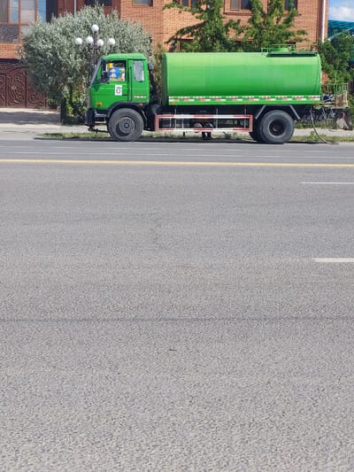 A bright green water truck is parked on the side of a street in Kyzylorda, Kazakhstan. The truck has a large cylindrical tank mounted on its chassis. In the cab of the truck, a driver can be seen. Behind the truck, a brick building and lush green trees are visible. The street is paved and has yellow and white lane markings.