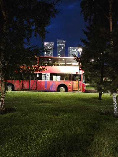 This media file features a double-decker bus parked on a grassy area at night, with a cityscape visible in the background. The bus is painted red and decorated with colorful abstract patterns. Tall, modern buildings with illuminated windows are scattered across the skyline, suggesting an urban setting. The sky is a deep blue, indicating it is nighttime. Trees frame the scene on both the left and right sides, their dark foliage contrasting with the illuminated bus and buildings. The grass in the foreground is lush and green, with some subtle lighting casting shadows. There are no people visible in the frame. The overall mood is tranquil, with the lights of the city and the bus creating a somewhat surreal or fantastical atmosphere. The presence of the bus in this setting could imply a temporary installation, a special event, or a unique form of public transport.