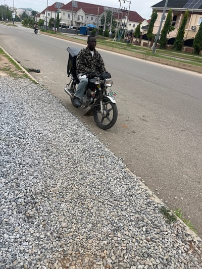 In Karum, Nigeria, a young man with dark skin is riding a motorcycle. He is wearing a light-colored shirt with a brown and black animal print pattern and blue jeans. The motorcycle has a black storage box mounted behind the seat. The scene is on a paved road with a gravel shoulder. In the background, there are houses with solar panels on their roofs and trees. Another person is riding a motorcycle in the distance. The sky is overcast.