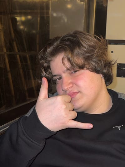 A young person with curly brown hair, fair skin, and freckles is positioned at an angle, facing the viewer with a playful expression. The individual is wearing a black sweatshirt with a gray accent at the shoulder. Their hand is making a "shaka" sign, with the thumb and pinky extended. A reflective surface is visible behind them, hinting at an indoor setting with some overhead lighting.