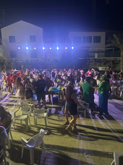 This image depicts a nighttime outdoor gathering, possibly a party or event, in Blanca, Spain. A crowd of approximately 30-40 people, mostly young adults, are present. The environment appears to be an open area with a paved ground, illuminated by stage lights that cast long shadows. In the background, there are buildings that suggest a residential or commercial setting. Several white plastic chairs are scattered around the area, some occupied and others empty. A makeshift table with drinks and other items is visible on the right side of the frame. One individual in the foreground is dressed in a green outfit with blue overalls, resembling a character costume. People are standing, conversing, and some appear to be dancing, suggesting a lively atmosphere. The overall scene is indicative of a social event taking place after dark.